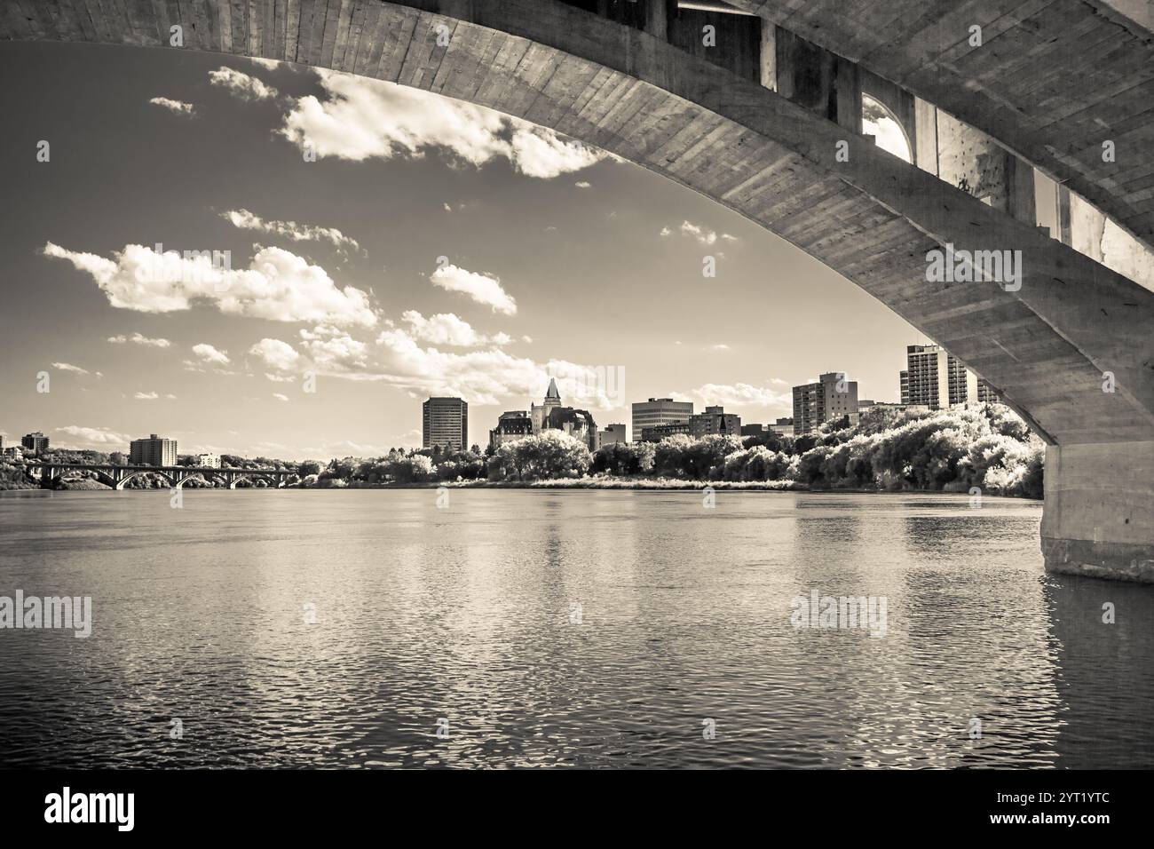 A city skyline is visible through a bridge over a river. The bridge is a large, arched structure that spans the width of the river. The city is bustli Stock Photo