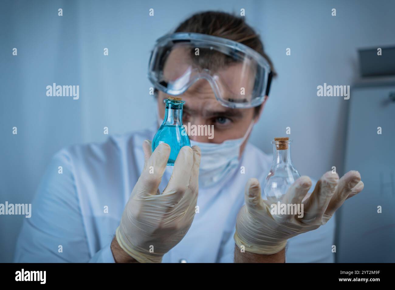 A scientist wearing protective goggles, gloves, and a face mask carefully analyzing vials containing blue and clear liquids, emphasizing precision, re Stock Photo