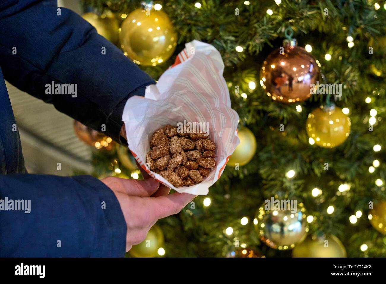 Augsburg, Bavaria, Germany - December 5, 2024: Man holding a bag of roasted almonds in front of a festively decorated Christmas tree, sweet treat at Christmas time *** Mann hält eine Tüte gebrannte Mandeln vor einem festlich geschmückten Weihnachtsbaum, süßer Genuss zur Weihnachtszeit Stock Photo