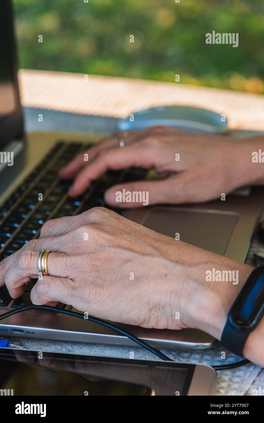 Close-up of elderly woman's hands typing on laptop keyboard, working remotely in outdoor setting Stock Photo