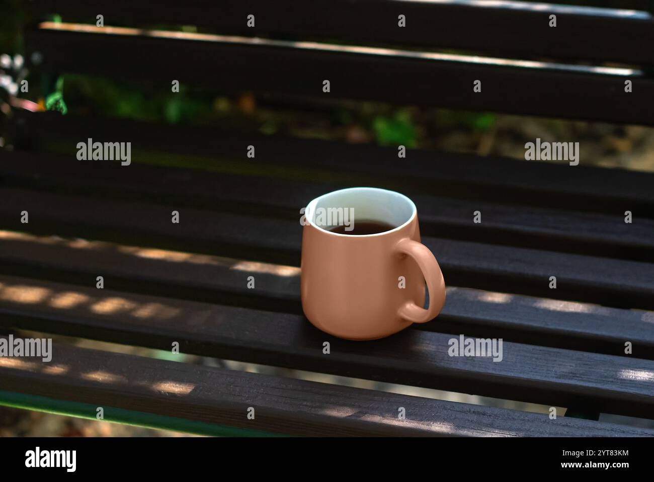 A coffee cup sits on a wooden bench in the shade. The cup is half full of coffee. Concept of relaxation and leisure. Mocha Mousse color of the year 20 Stock Photo