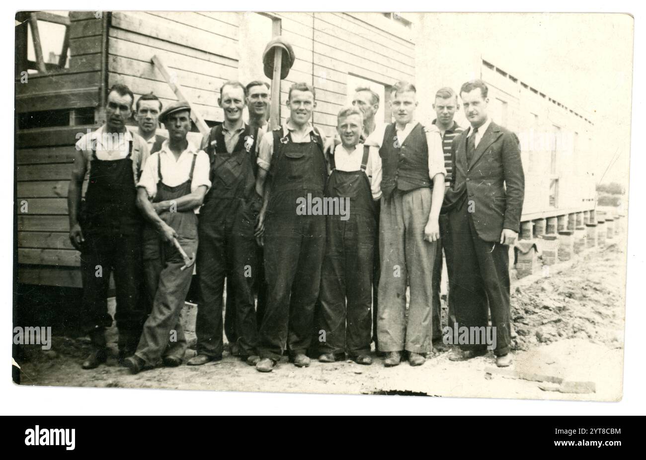 Original 1920's era postcard of working class men - builders or navvies, constructing wooden huts, holding tools, maybe the Foreman standing wearing a suit and tie at the end of the line. Some men wearing flat caps, late 1920's or early 1930's U.K. Stock Photo