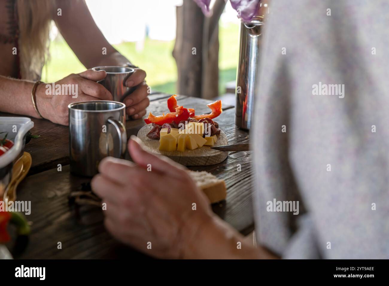 Wooden board with cheese, sausage and peppers stands on a rustic table between people Stock Photo