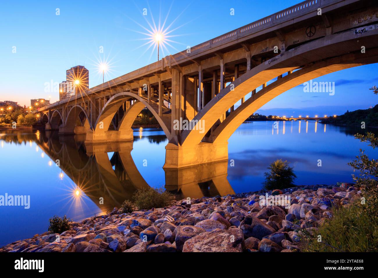 A bridge spans a river at dusk. The bridge is lit up with lights, creating a serene and peaceful atmosphere. The reflection of the lights on the water Stock Photo