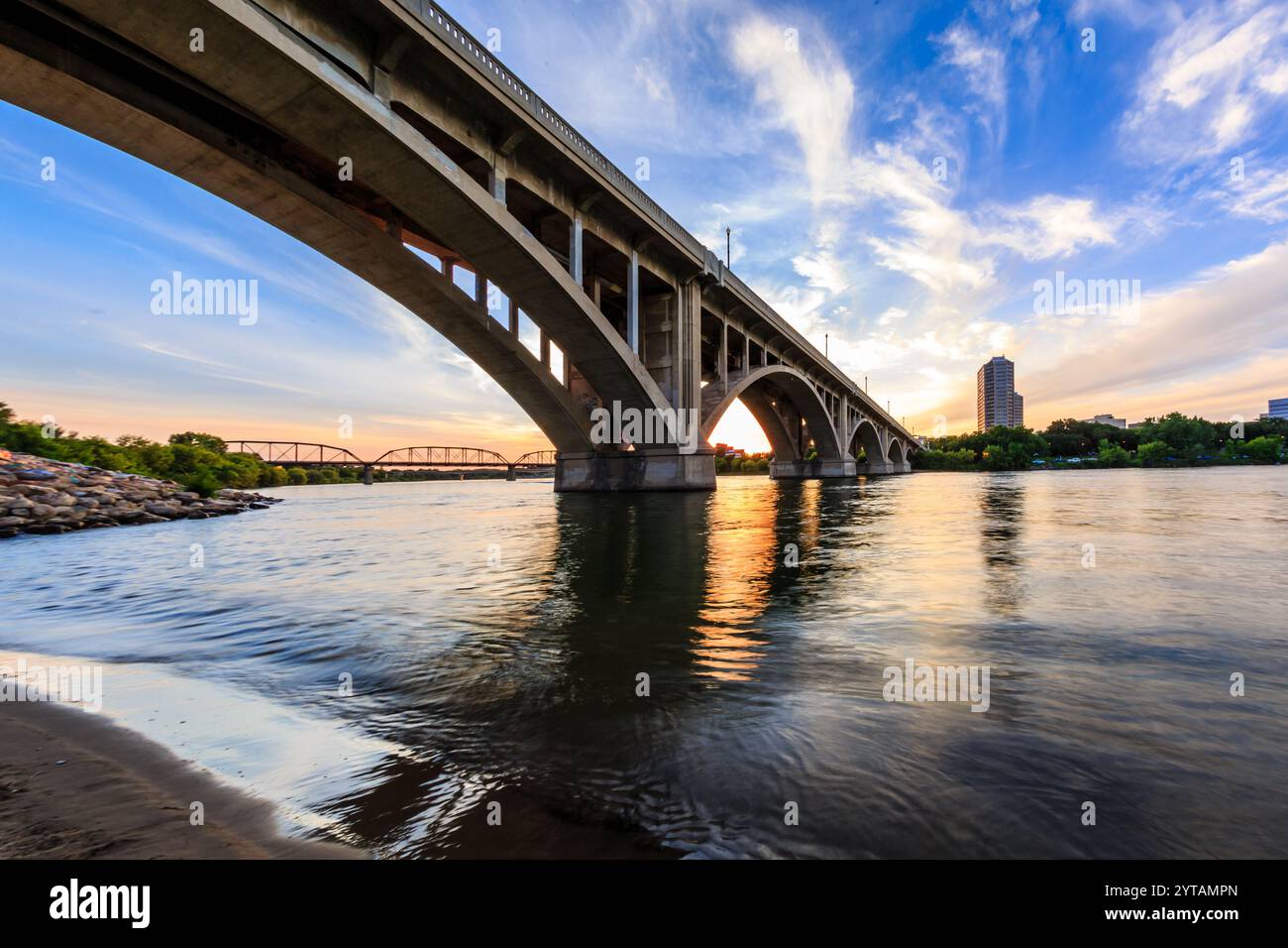 A bridge spans a river with a beautiful sunset in the background. The bridge is a large structure that connects two sides of the river Stock Photo
