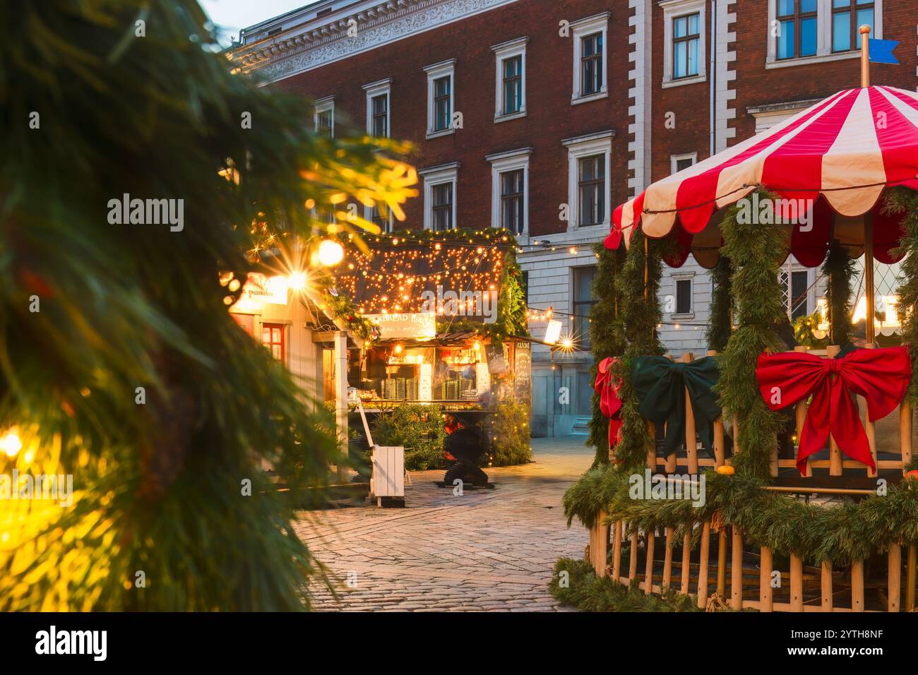 Dome square Christmas market scene with decorated red-and-white stalls, pine garlands, fairy lights, and a gazebo wrapped in greenery and bows, creati Stock Photo