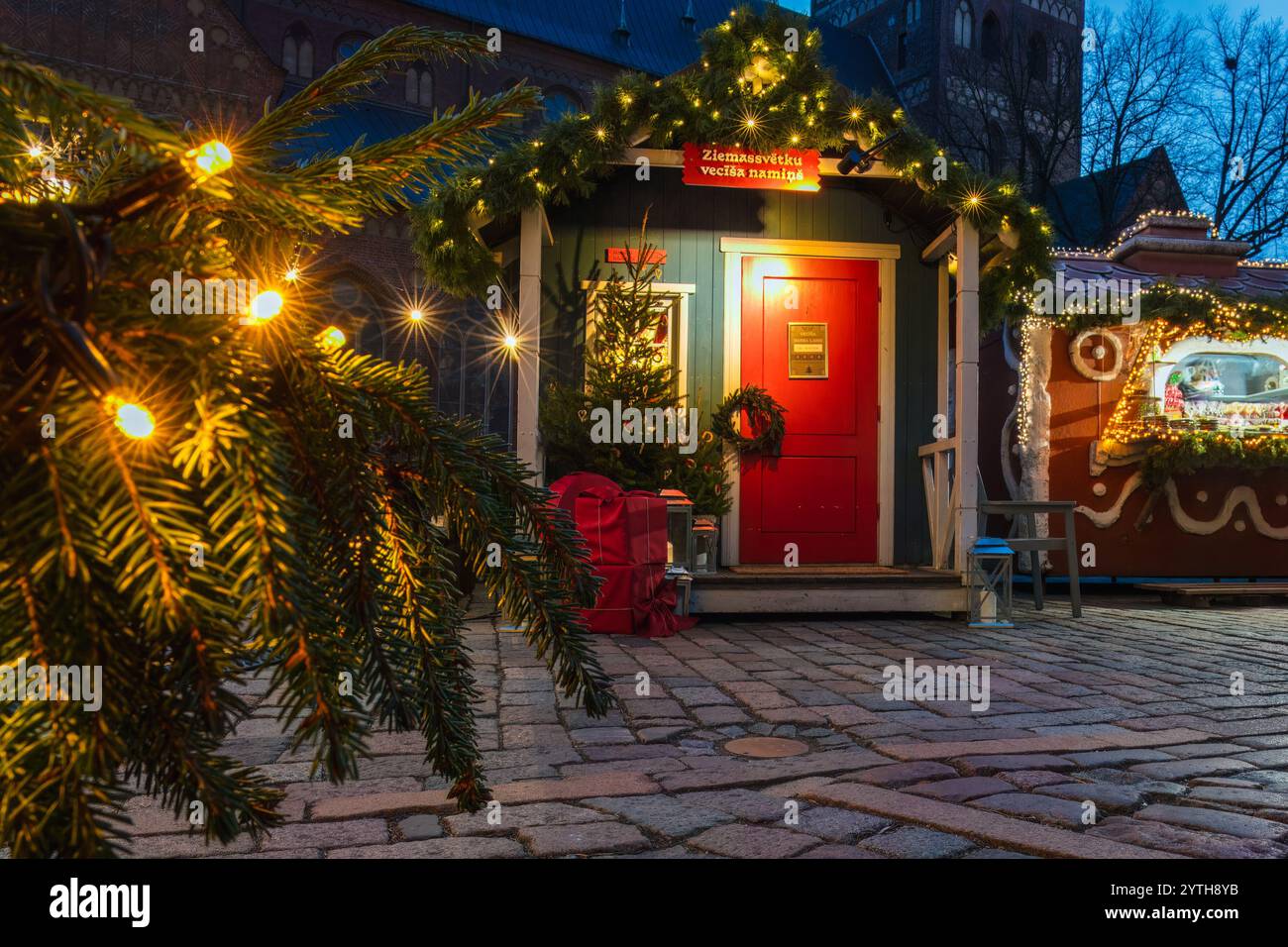 Festive Santa Claus cabin with a bright red door, garland decorations, and Christmas tree surrounded by presents, illuminated by warm fairy lights on Stock Photo