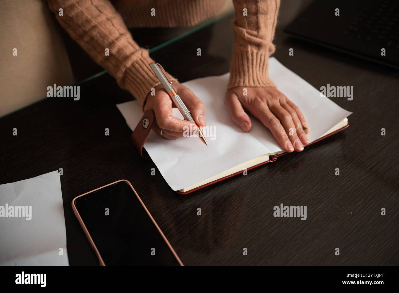 A woman in a sweater writing notes in a notebook. A smartphone and laptop are visible on the desk, suggesting a productive work or study session. Stock Photo