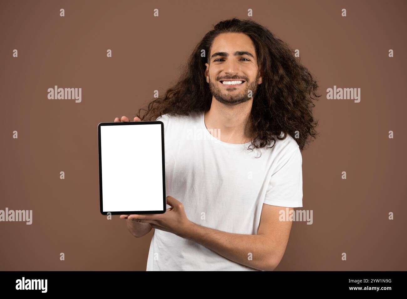 Young man with curly hair joyfully presenting tablet against neutral backdrop Stock Photo