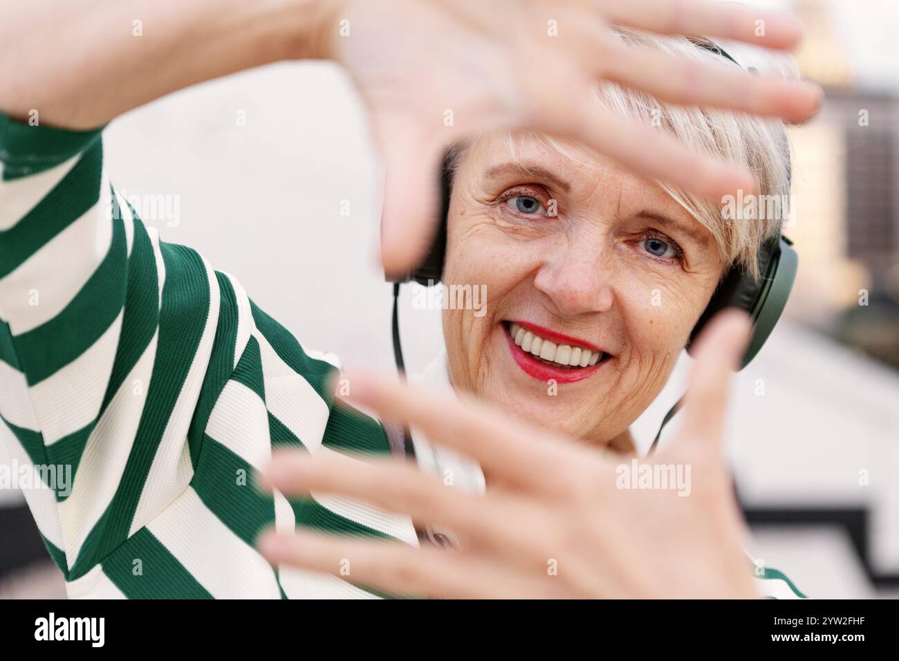 Joyful Senior Woman Making Frame with her Hands While Listening to Music with Headphones, Smiling and Looking at Camera. Stock Photo