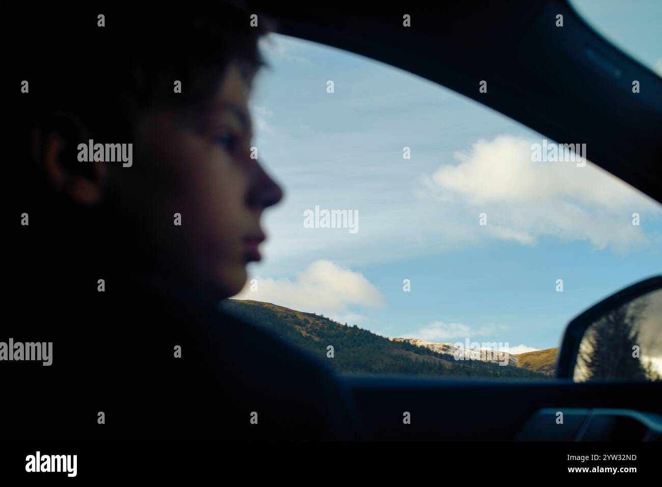 Close-up side profile of a child looking out a car window with a scenic view of mountains and blue sky in the background, Highlands, Scotland Stock Photo