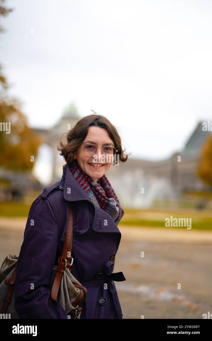 Smiling woman in a purple coat with a scarf standing before a historical monument on a cloudy day. Stock Photo