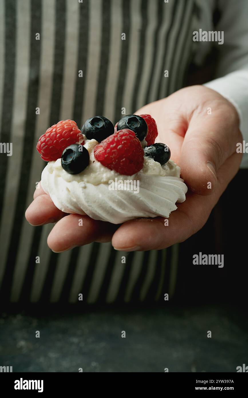 Close-up of a hand holding a mini meringue dessert topped with whipped cream, fresh blueberries, and raspberries against a striped fabric background. Stock Photo