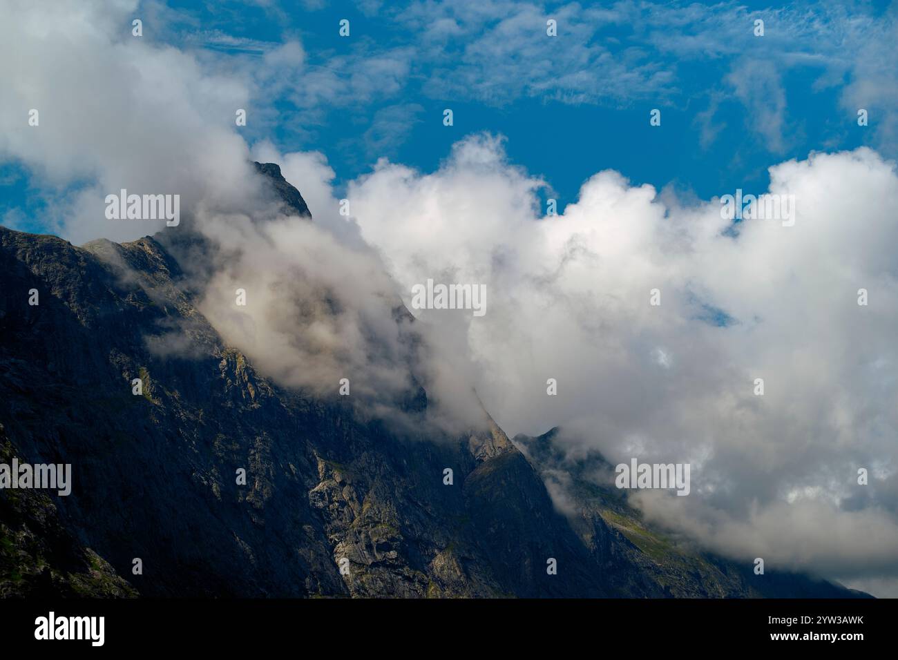 Clouds shroud the rugged peaks of a mountain under a bright blue sky ...