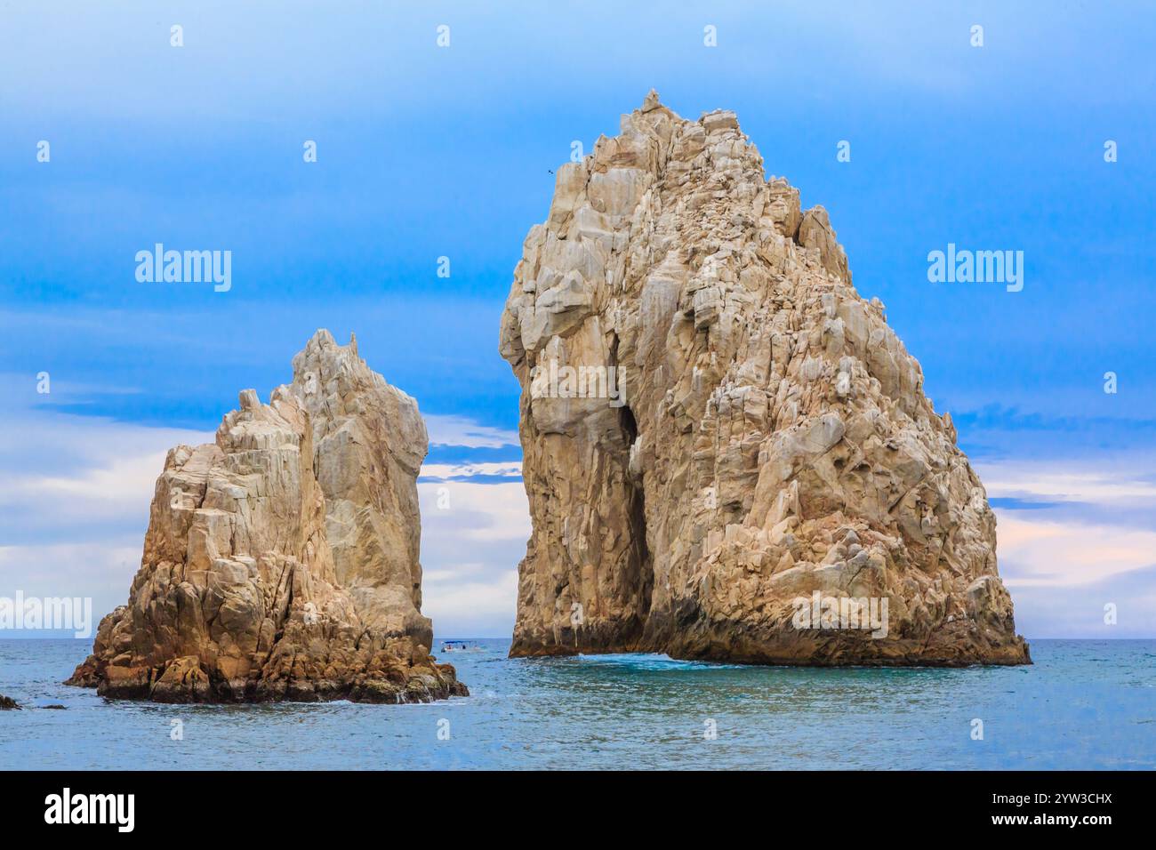 A large rock formation sits on the ocean shore. The sky is blue and ...