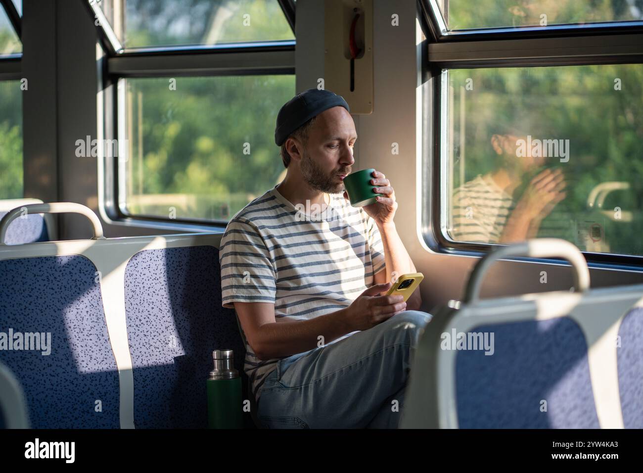 Calm man sip tea from thermos, chatting on smartphone in train car in morning. Male travel pause Stock Photo