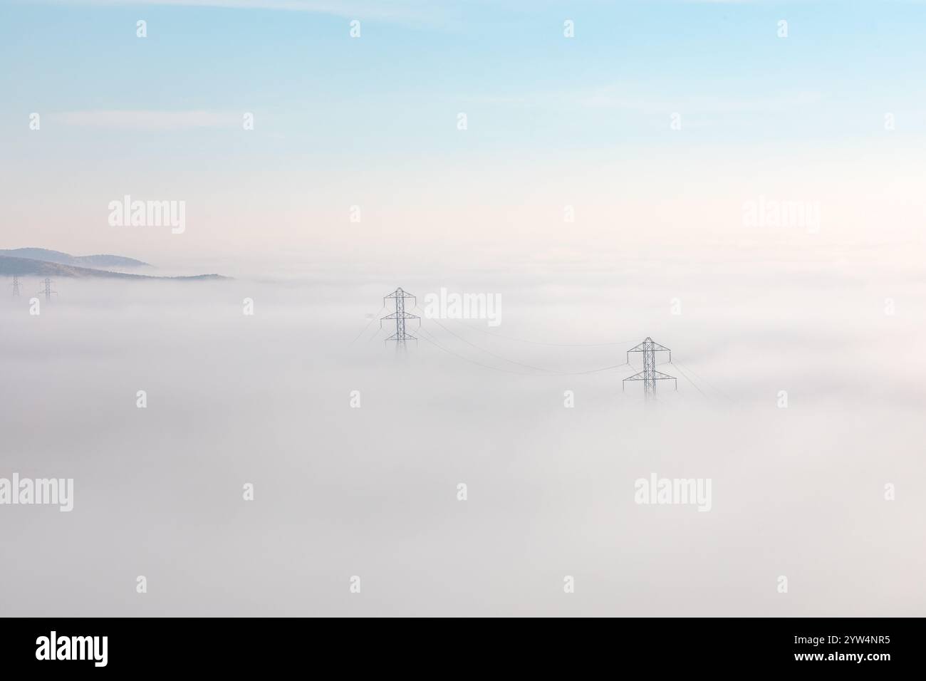 A landscape view of power lines stretching across a thick layer of fog. The fog obscures the terrain, with only the tops of hills visible in the dista Stock Photo