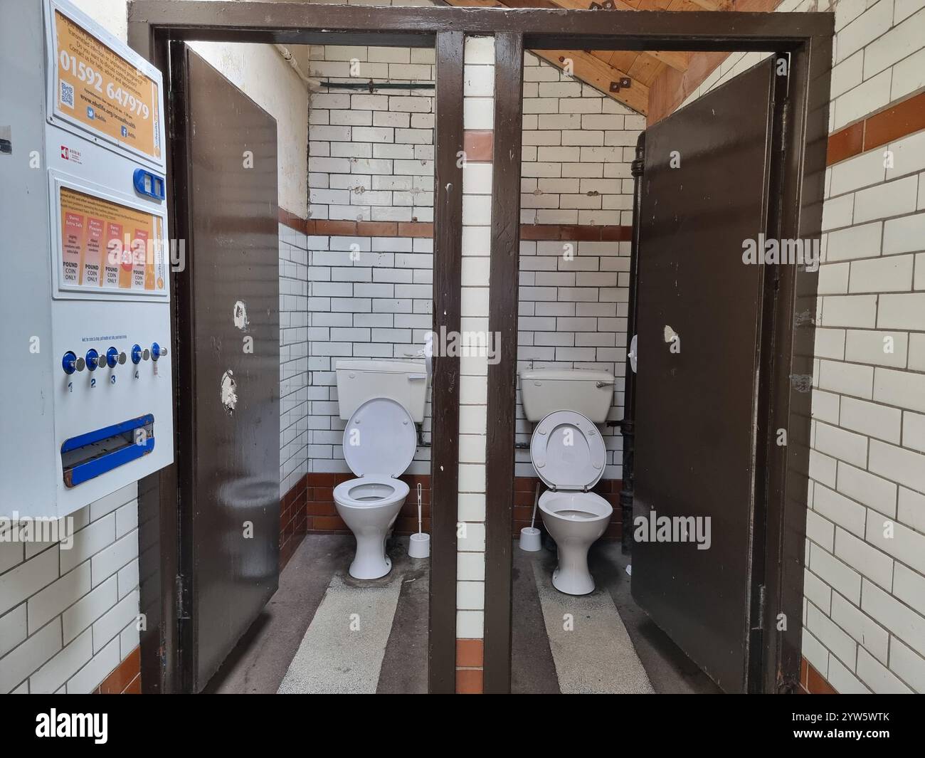 View on two white public toilets, with toilet brushes through open doors; a vending machine on the wall Stock Photo