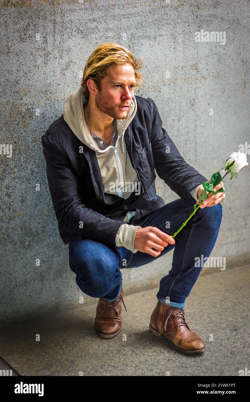 A man with blond hair crouches against a concrete wall, holding a white ...