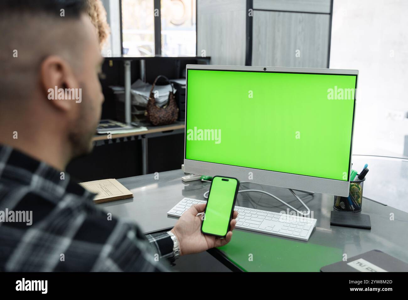 Businessman working at a modern office desk, using a computer and smartphone with a green screen for creating a mockup display Stock Photo