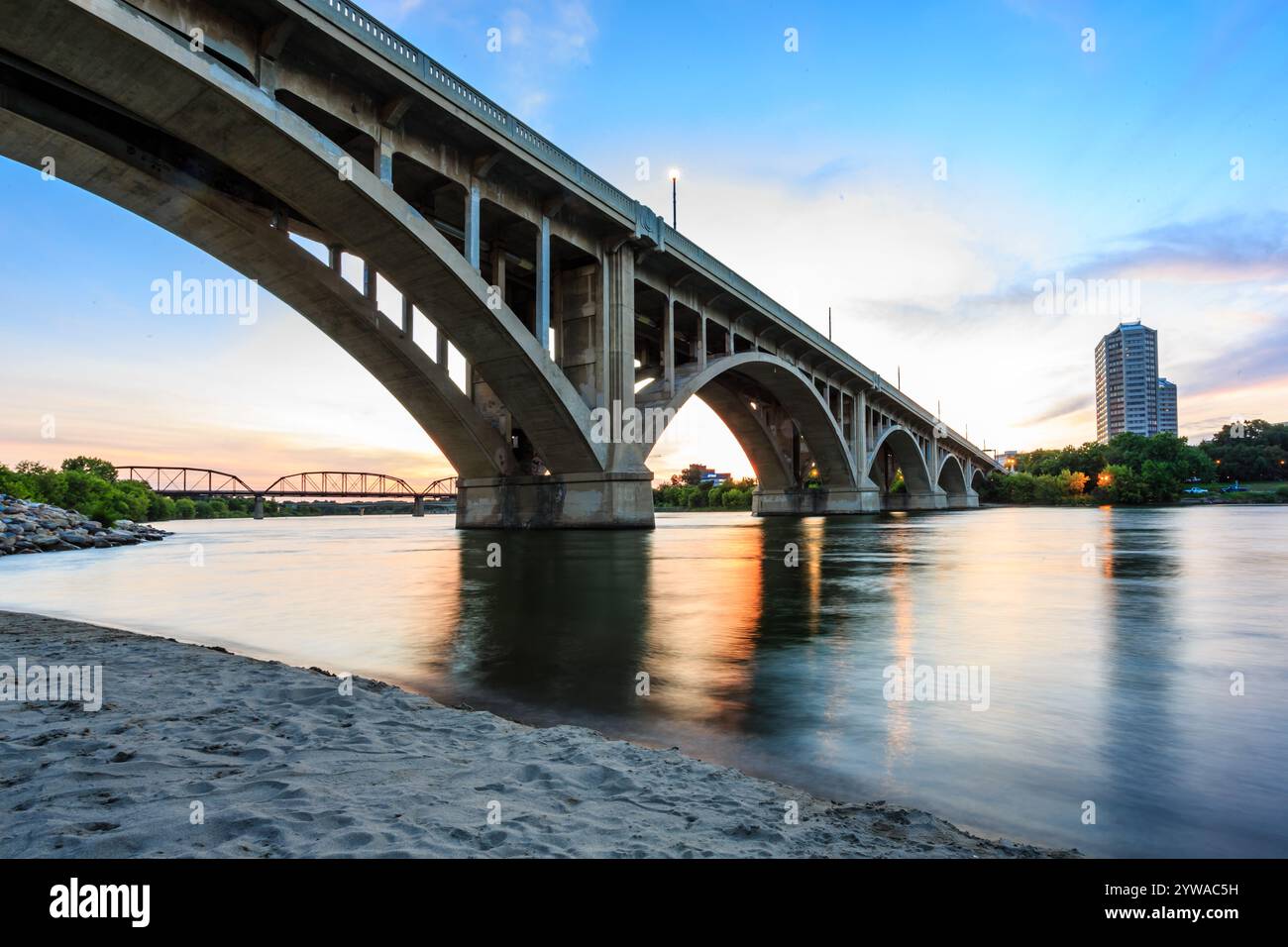 A bridge spans a river with a city in the background. The bridge is illuminated by lights, creating a serene and peaceful atmosphere Stock Photo