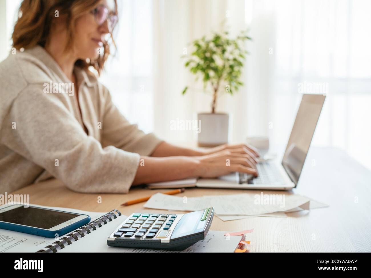 Beautiful middle-aged woman in glasses making marks in financial documents at home office using laptop. Small business, home finances, money savings c Stock Photo