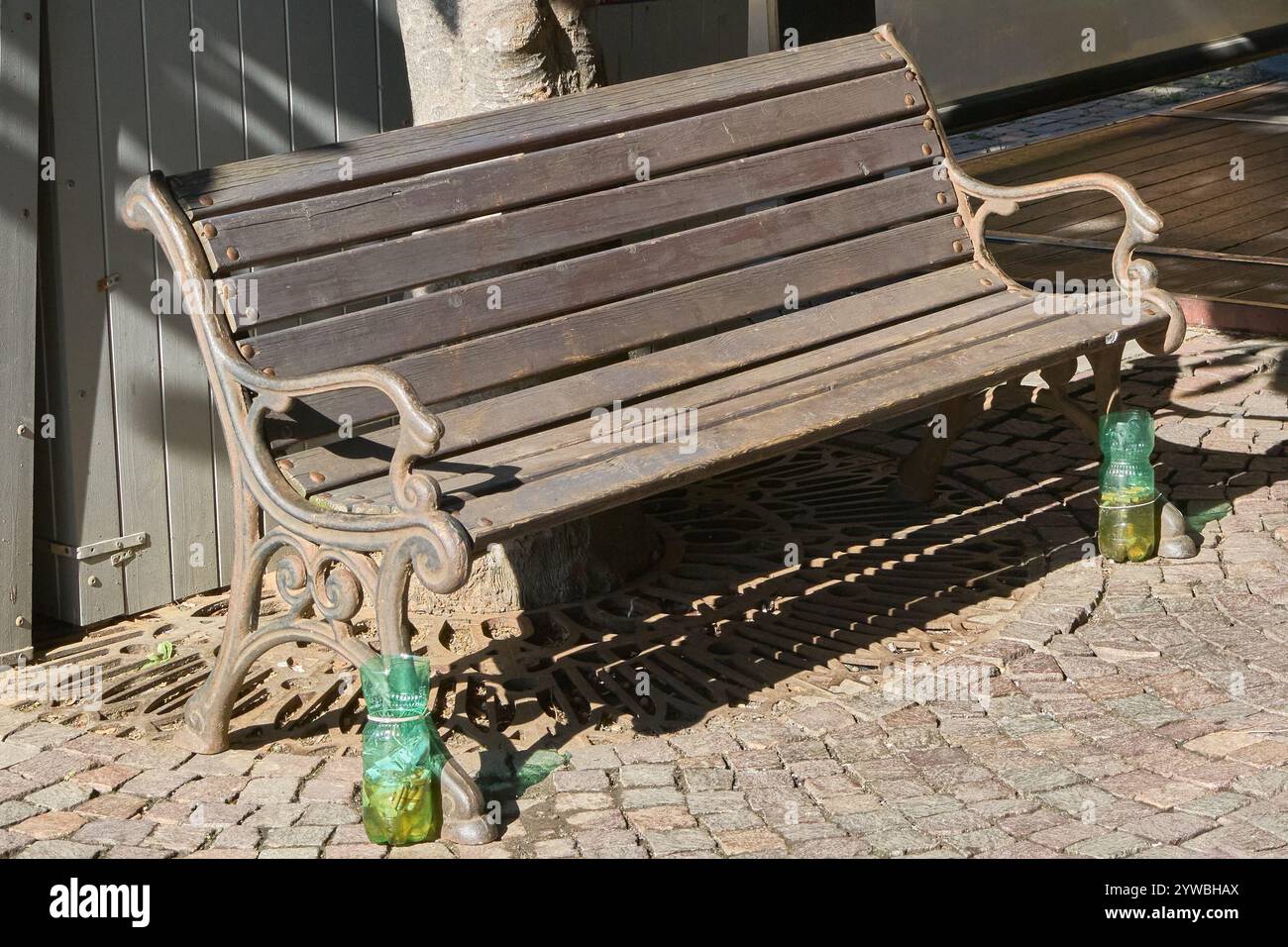 A wooden bench with wrought iron structure located on a cobblestone street, featuring recycled green plastic bottles used as ashtrays at each end. The Stock Photo