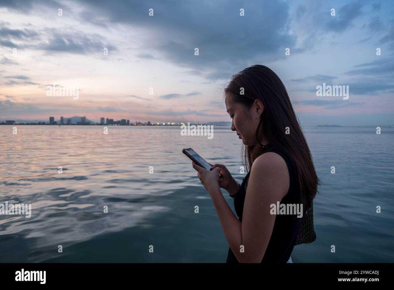 Side view of a young woman standing by the sea using her mobile phone, Tan Jetty, George Town, Penang, Malaysia Stock Photo
