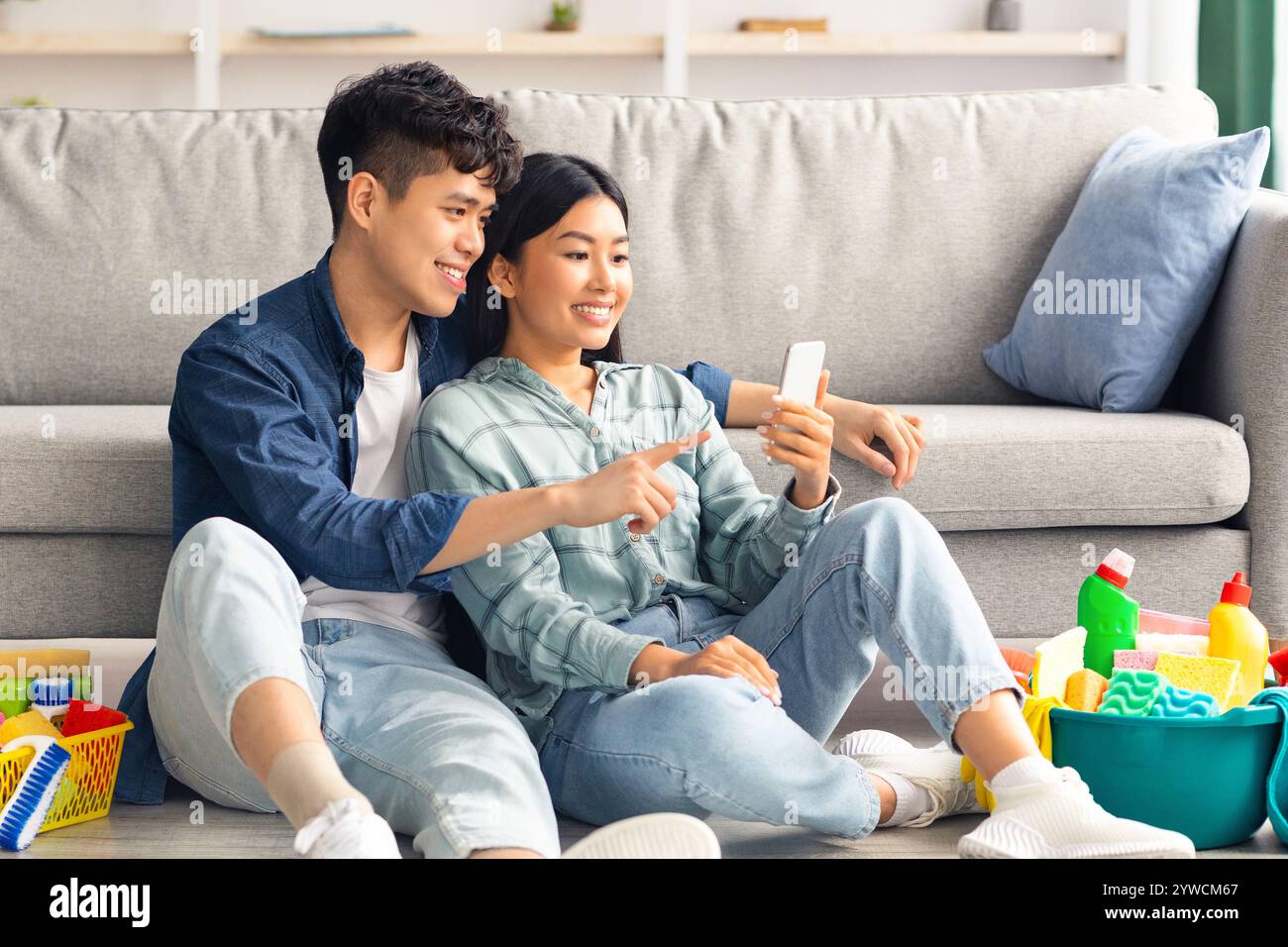 Loving chinese couple with cleaning tools resting, using smartphone Stock Photo