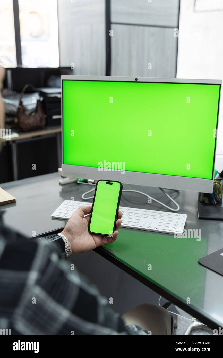 Office worker holding a smartphone with a vibrant green screen, positioned near a computer featuring a matching green screen on the desk Stock Photo