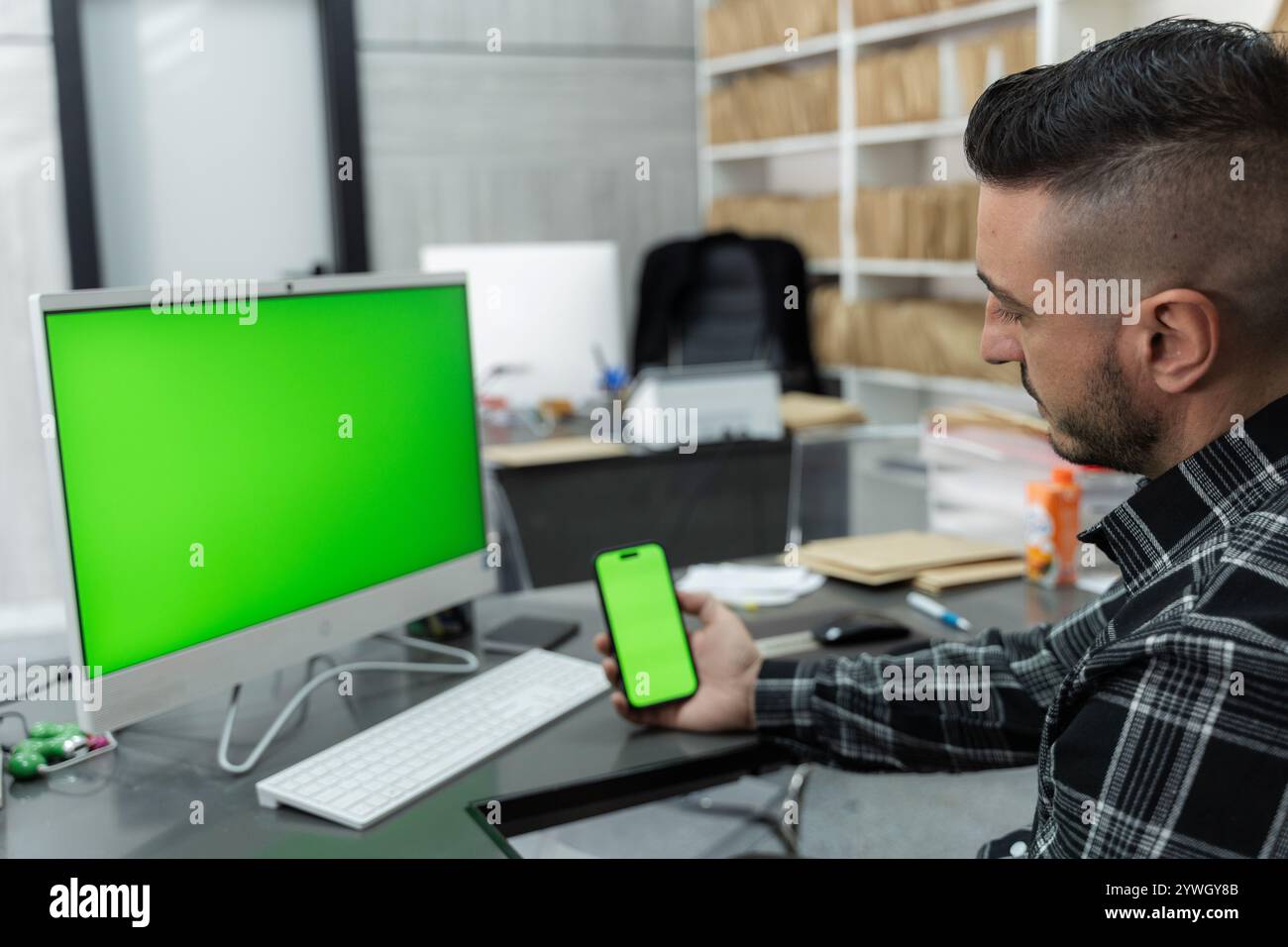 Businessman sitting at desk comparing green screen on computer monitor and smartphone in modern office Stock Photo