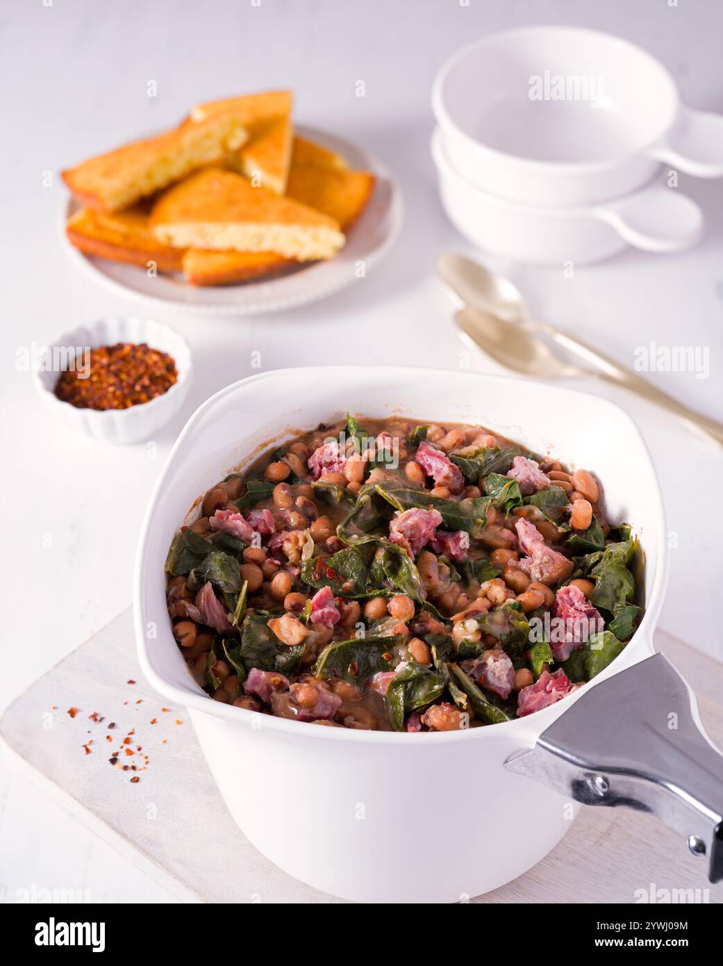 Black-eyed peas with collard greens and ham hocks in a large white cooking pot with corn bread on the side Stock Photo