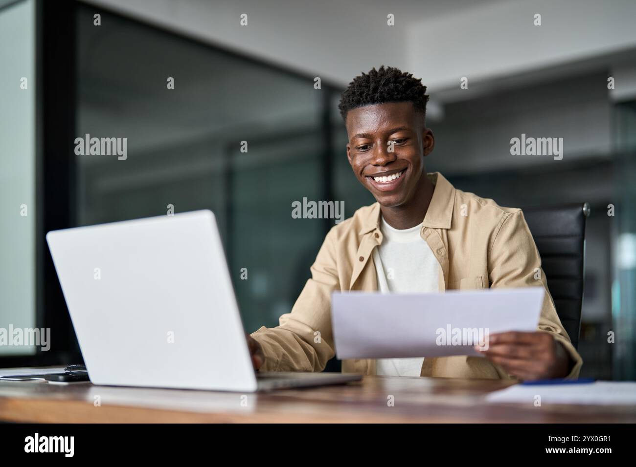 Happy young African business man holding documents using laptop at work. Stock Photo