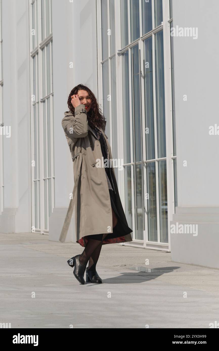 Woman with curly hair, dressed in a trench coat, walking along a white ...