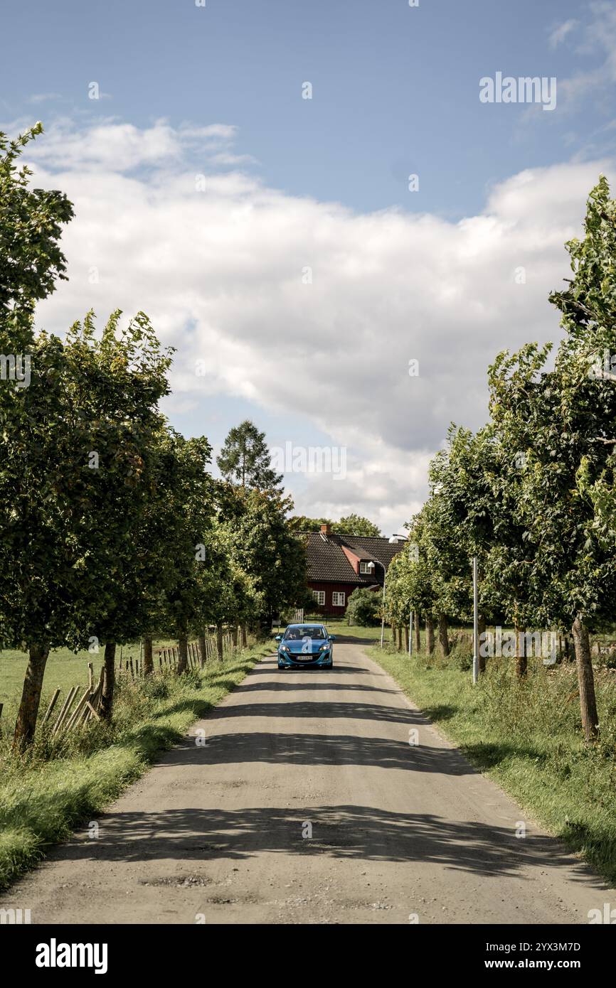 Blue car driving on rural road lined with trees and traditional house ...