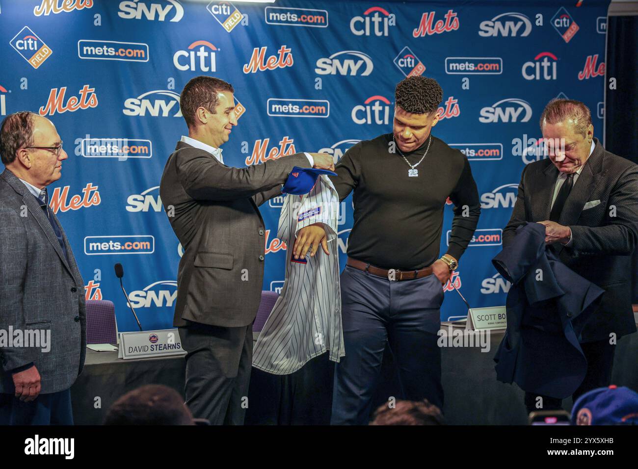 New York Mets outfielder Juan Soto #22 tries on his new Mets jersey after a press conference after being introduced at Citi Field in Corona, N.Y., Thursday, Dec. 12, 2024. Soto's 15-year, $765 million contract is the largest pact in  Major League Baseball history. Mets owner, chairman and CEO Steve Cohen, President of Baseball Operations David Stearns and Soto’s agent Scott Boras look on.(Photo: Gordon Donovan) Stock Photo