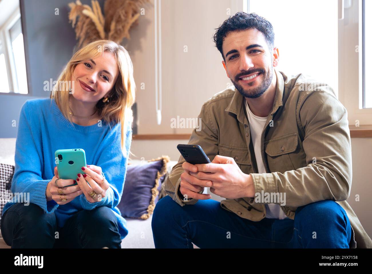 Couple sitting at home using smartphones, smiling and relaxed looking at camera Stock Photo