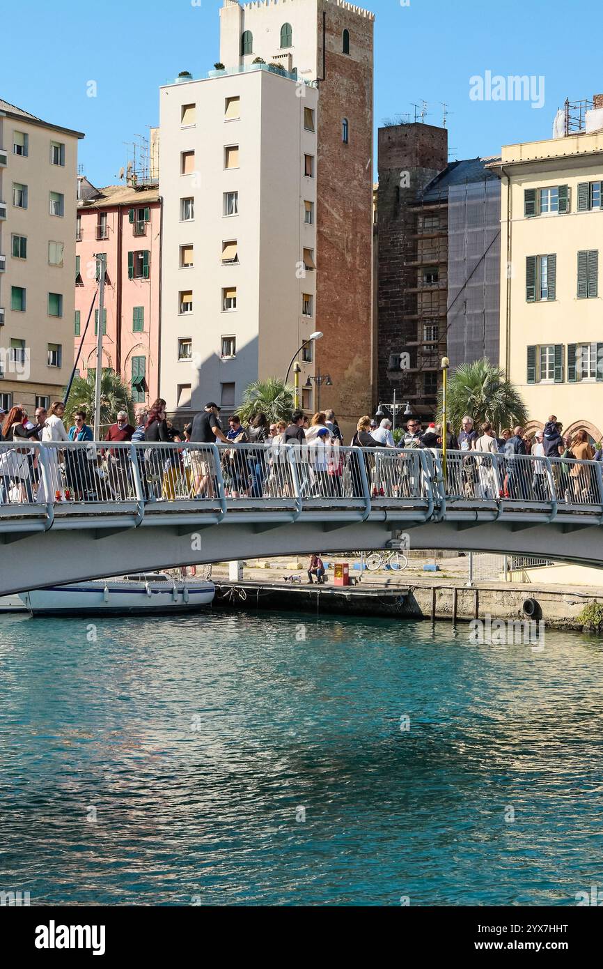 Savona, Italy, December 14, 2024: A vibrant pedestrian bridge bustling with tourists and locals under a sunny sky. Colorful buildings and a calm water Stock Photo