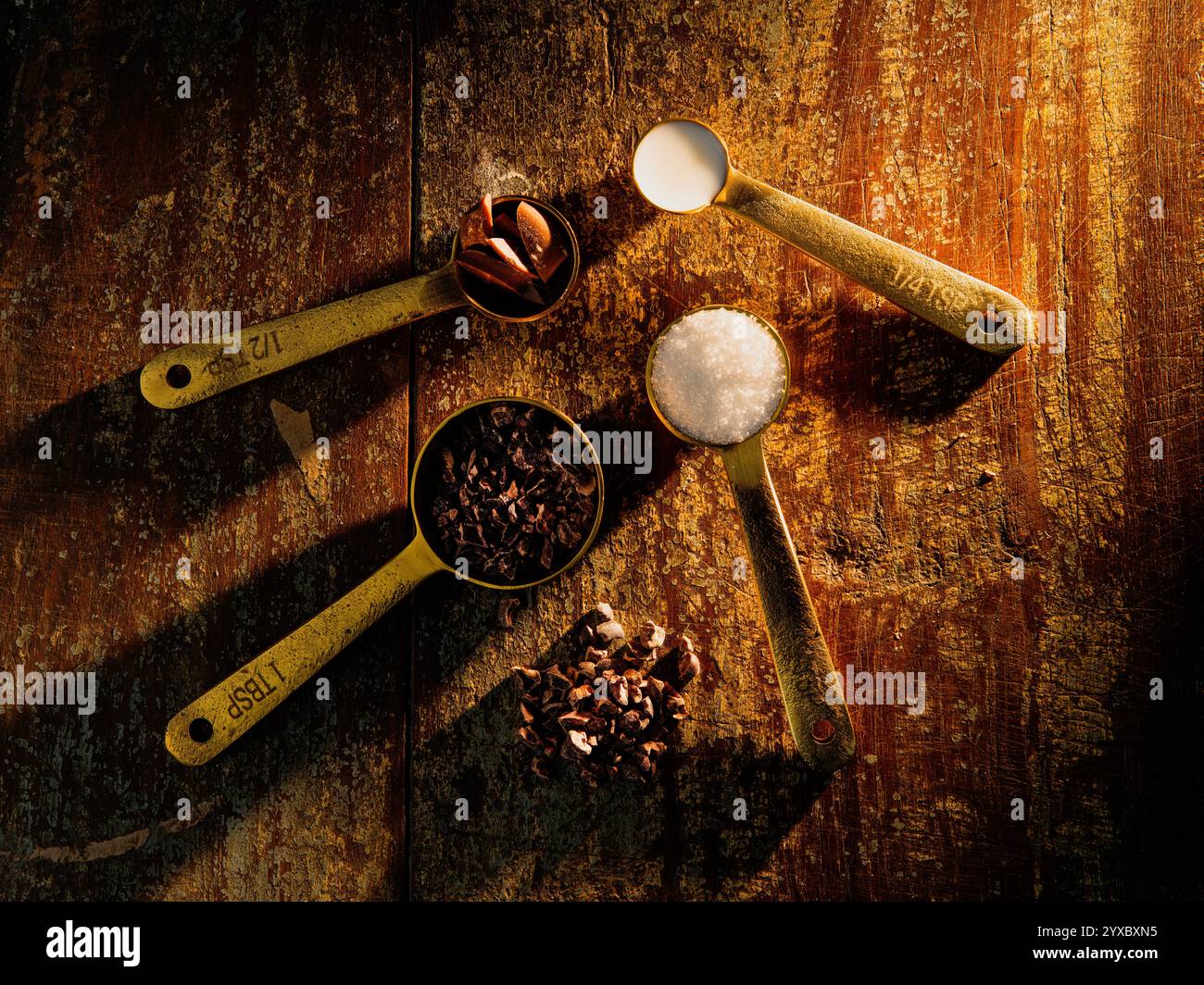 Golden Measuring Spoons with Ingredients Chocolate Cocoa Sugar Milk on distressed wooden backdrop dramatic lighting Stock Photo