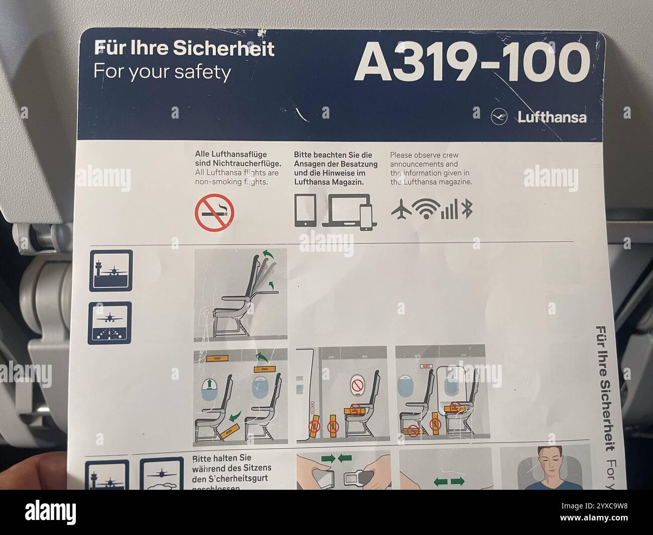 A Lufthansa A319-100 safety instruction card with non-smoking regulations, seat adjustment visuals, and emergency procedures, featuring diagrams and m - Smartphone Captured Stock Image