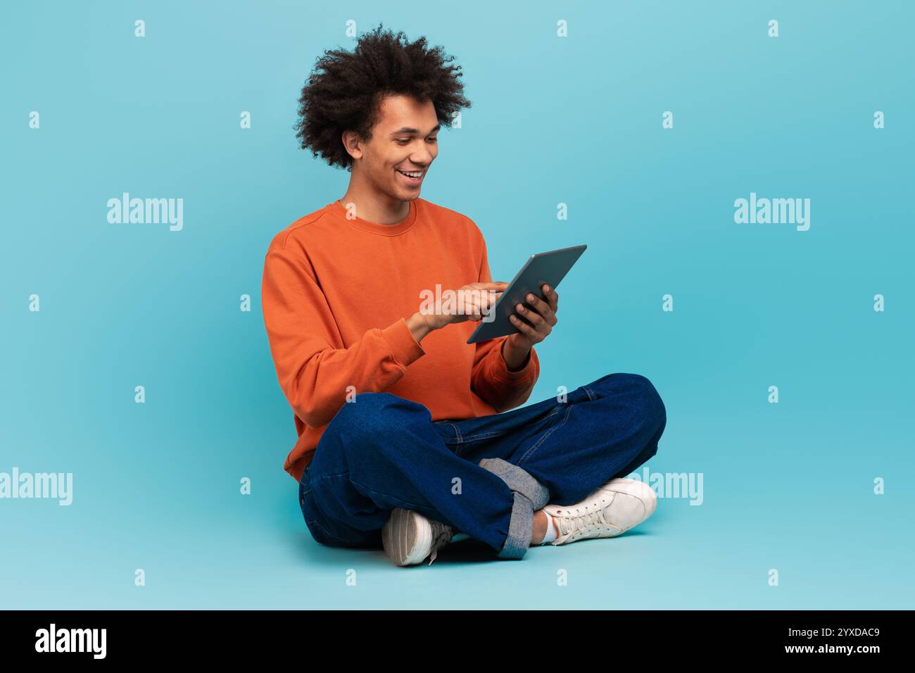 A young African American man sits cross-legged, smiling while using a tablet Stock Photo