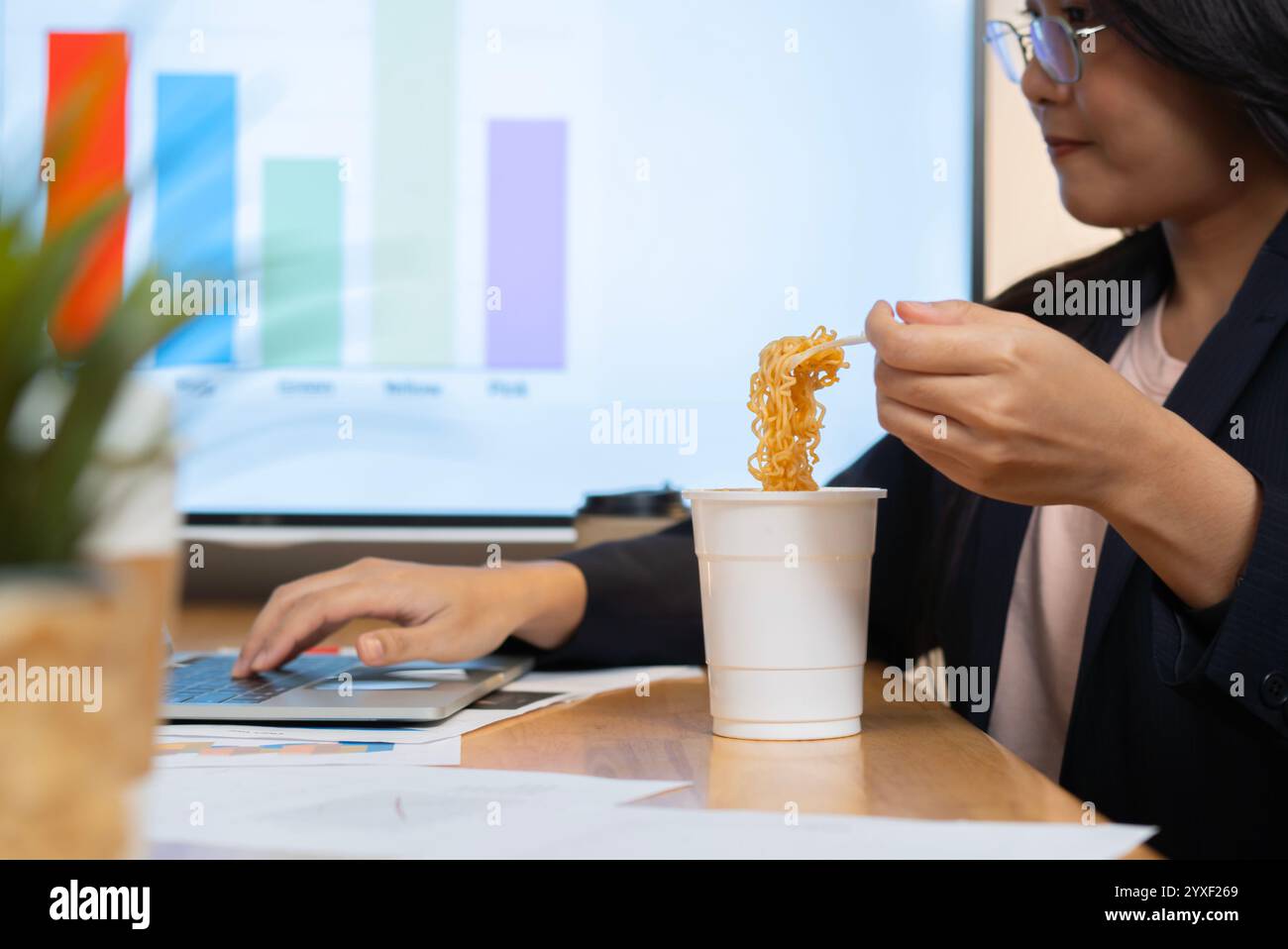 Happy Asian businesswoman has to eat noodles while working on laptop at office. Side view, selective focus. Stock Photo