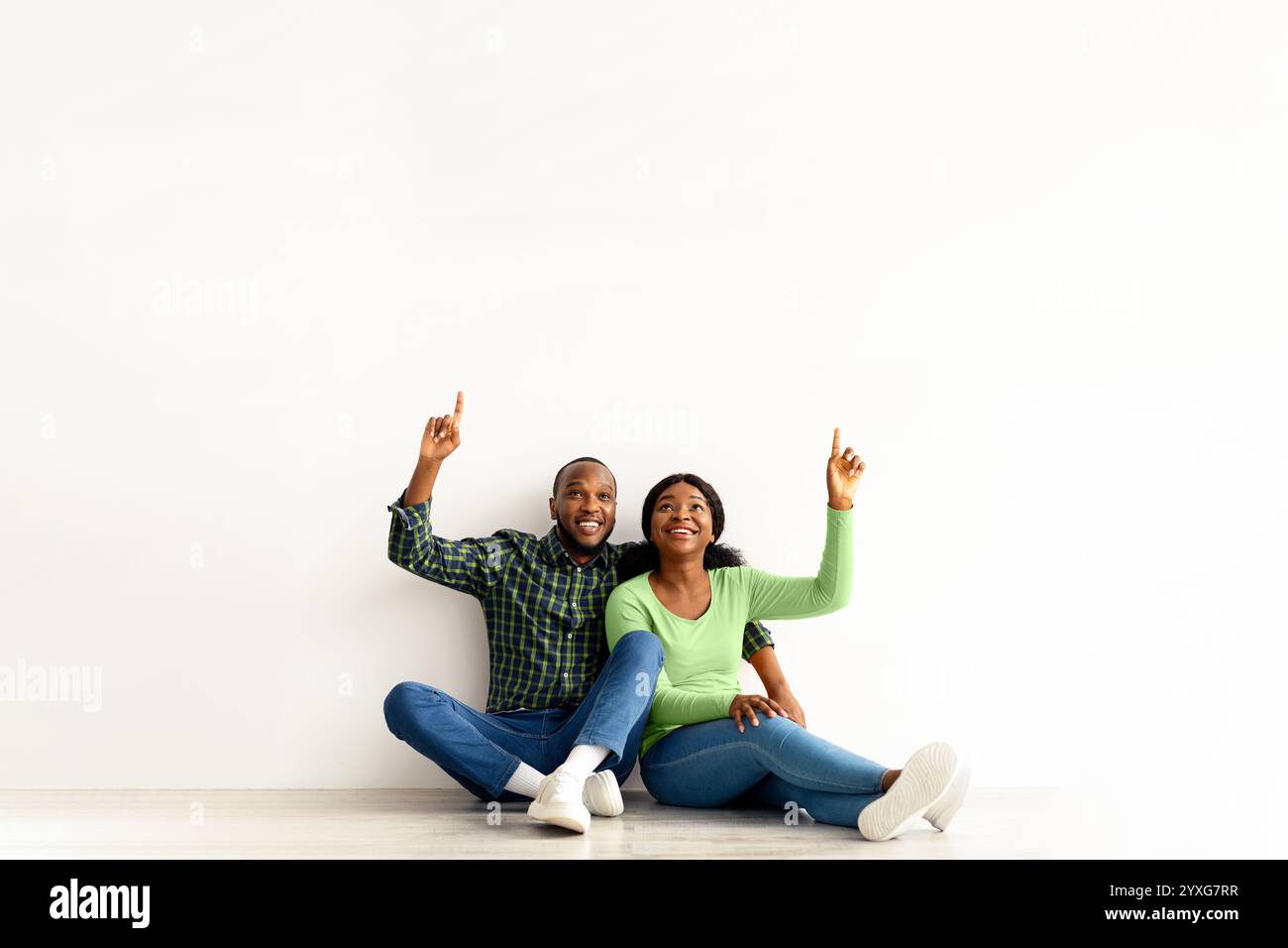 Happy African Couple Sitting On Floor Near White Wall And Pointing Up ...