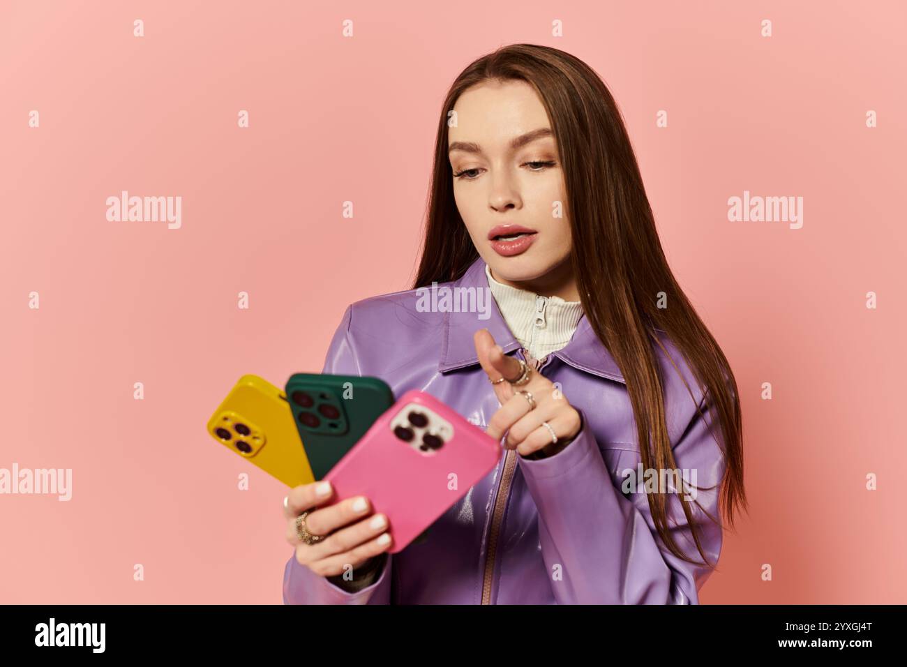 Stylish woman with colorful phone cases, interacting with her phone against a pink backdrop. Stock Photo