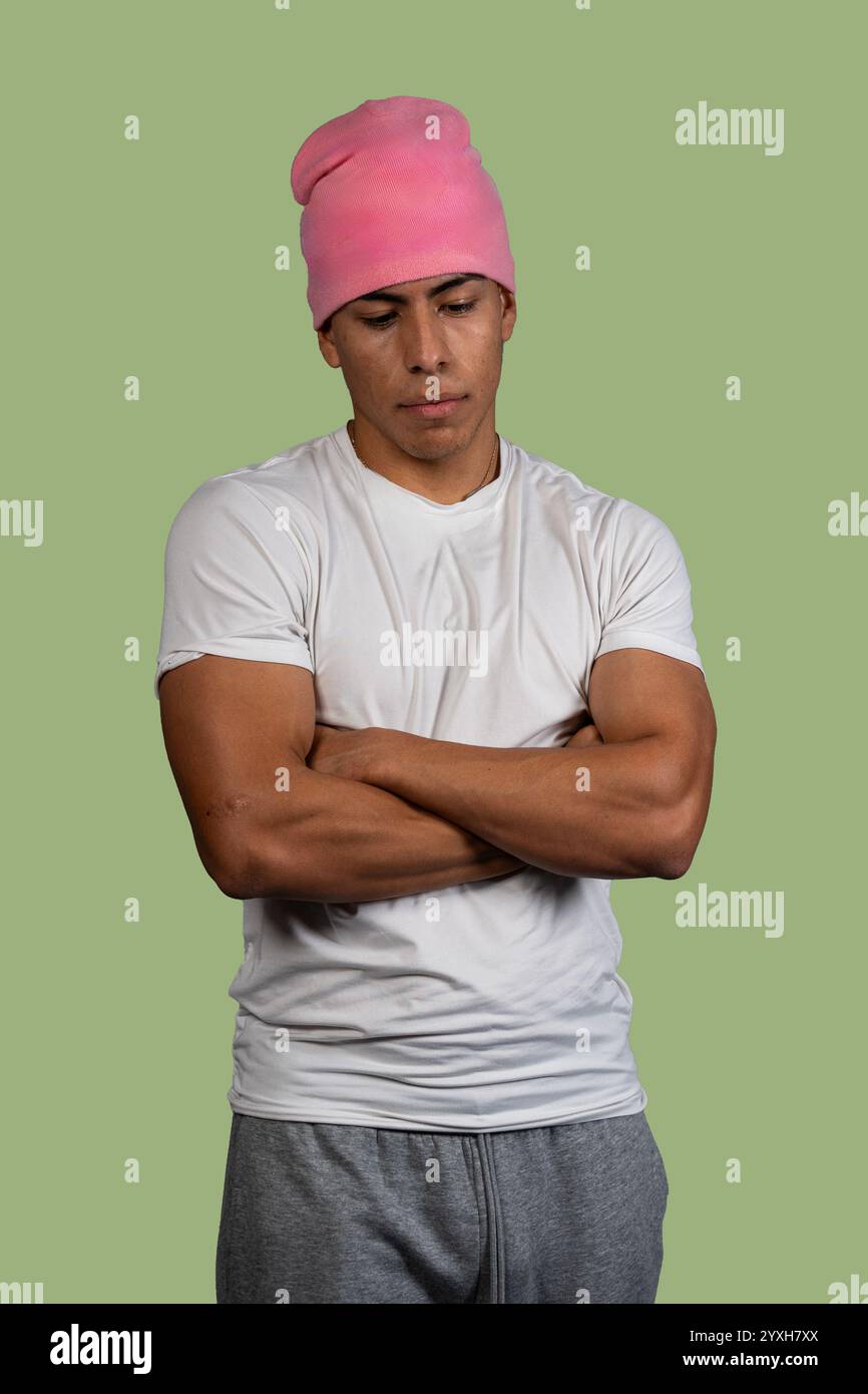 Studio portrait of a serious young latino man wearing pink beanie, white t-shirt and gray sweatpants, crossing his arms and looking down Stock Photo
