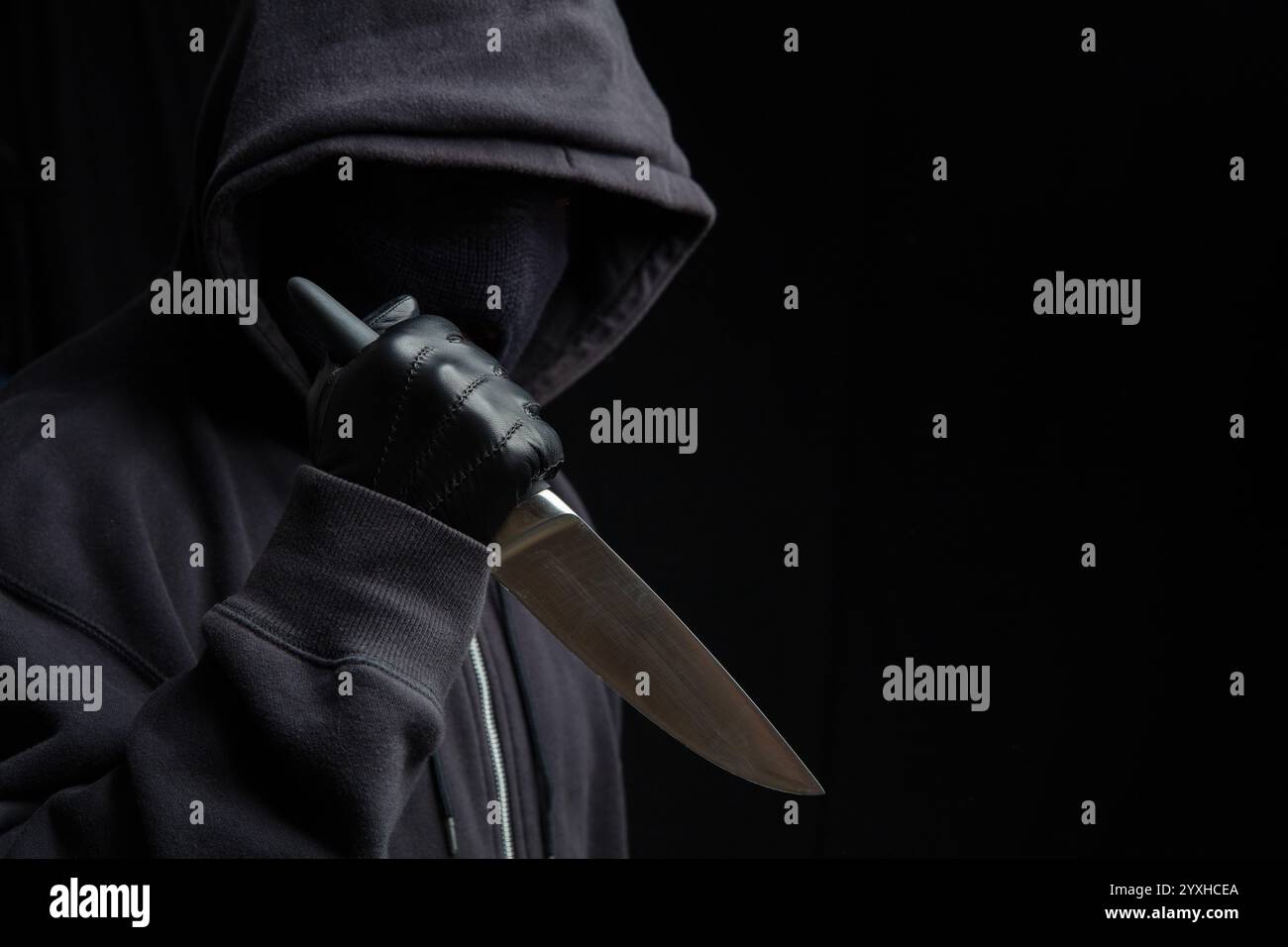 Criminal holding a knife, hooded man holding a weapon in gloved hand, black background, armed attack concept. Stock Photo