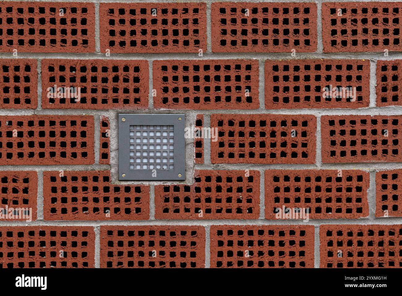 A textured brick wall features hollow red bricks arranged in a pattern, with a square light fixture mounted in the center, offering a modern design touch. Stock Photo