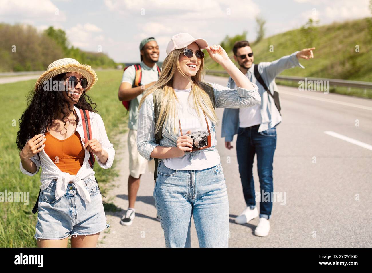 Group of millennial multiracial friends with backpacks walking along road, traveling together outdoors Stock Photo