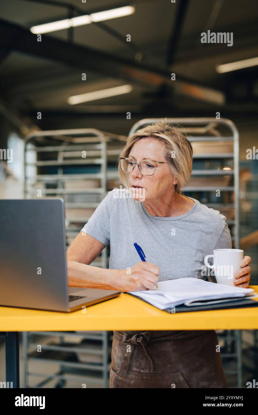 Focused senior female entrepreneur taking down notes while working at bakery Stock Photo