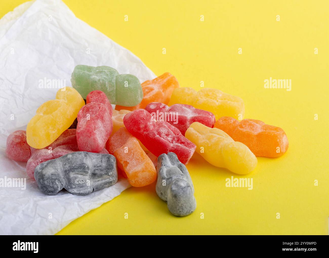 pile of vibrant jelly baby candies in red, green, orange, yellow, and black rests on crinkled white paper against a bright yellow background, Stock Photo